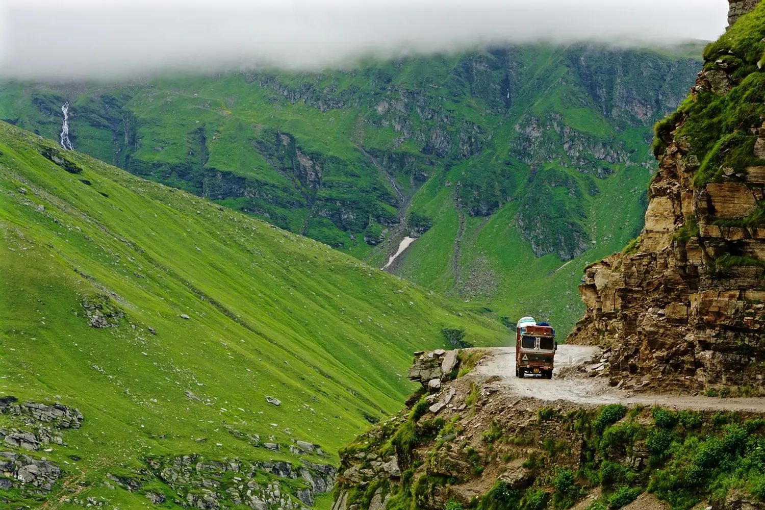 a truck passing through a narrow road near rohtang pass with green mountains in the backdrop on a cloudy day